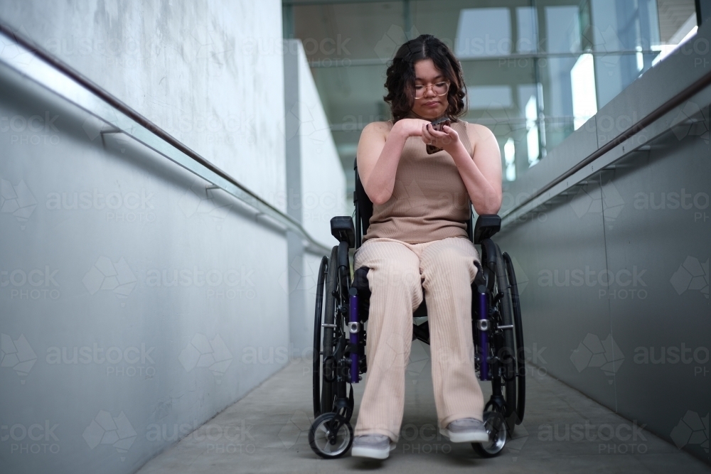 Smiling woman with a disability sitting in a wheelchair looking down at her mobile phone - Australian Stock Image