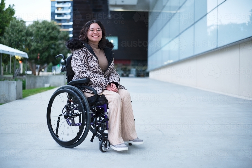 Smiling woman with a disability sitting in a wheelchair outside in a city - Australian Stock Image