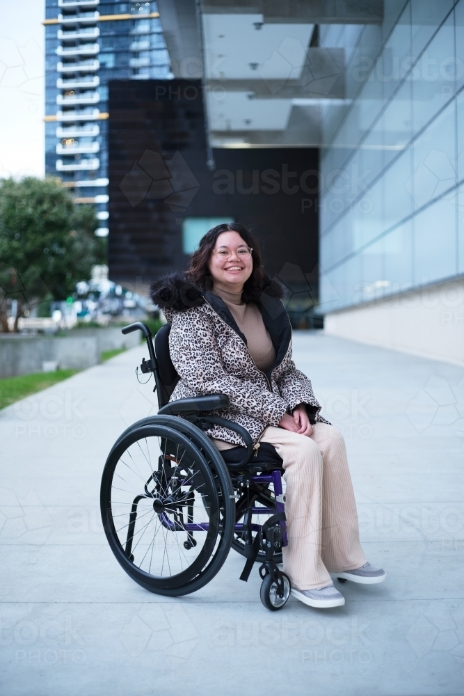 Smiling woman with a disability sitting in a wheelchair outside in a city - Australian Stock Image