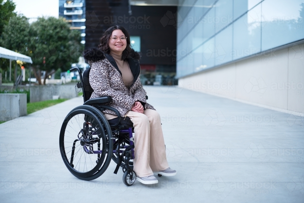 Smiling woman with a disability sitting in a wheelchair outside in a city - Australian Stock Image