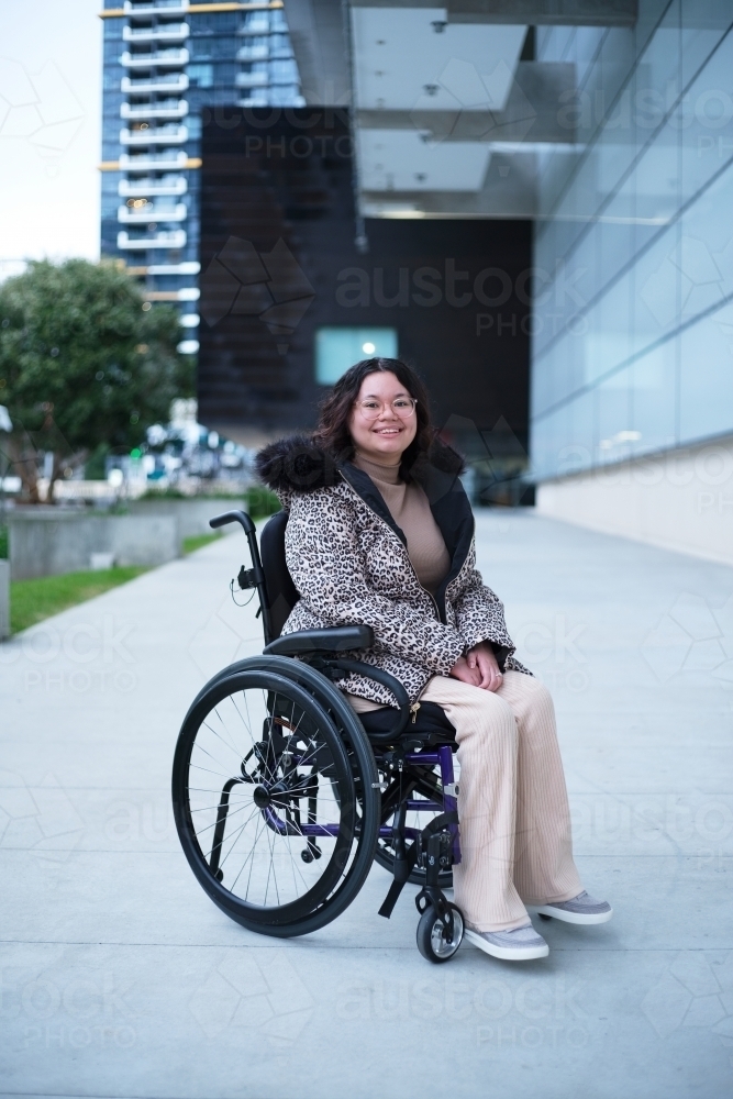 Smiling woman with a disability sitting in a wheelchair outside in a city - Australian Stock Image