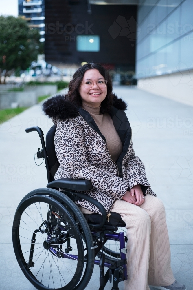 Smiling woman with a disability sitting in a wheelchair outside in a city - Australian Stock Image