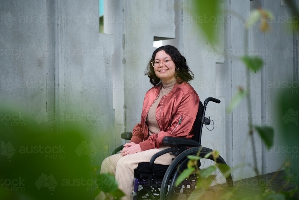 Smiling woman with a disability sitting in a wheelchair outside in urban setting with greenery - Australian Stock Image