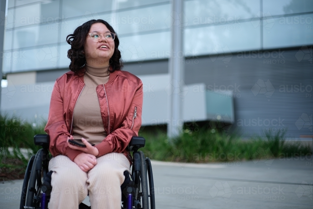 Smiling woman with a disability sitting in a wheelchair outside looking away from the camera - Australian Stock Image