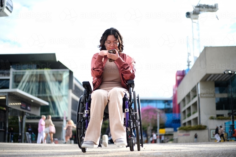 Smiling woman with a disability sitting in a wheelchair outside the city with mobile phone - Australian Stock Image