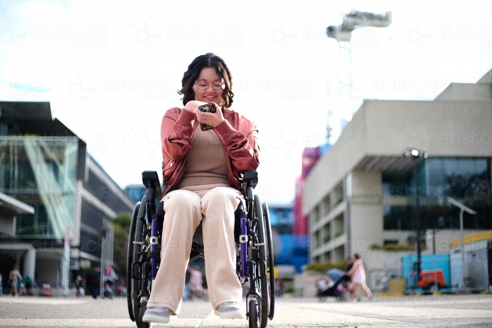 Smiling woman with a disability sitting in a wheelchair outside the city with mobile phone - Australian Stock Image