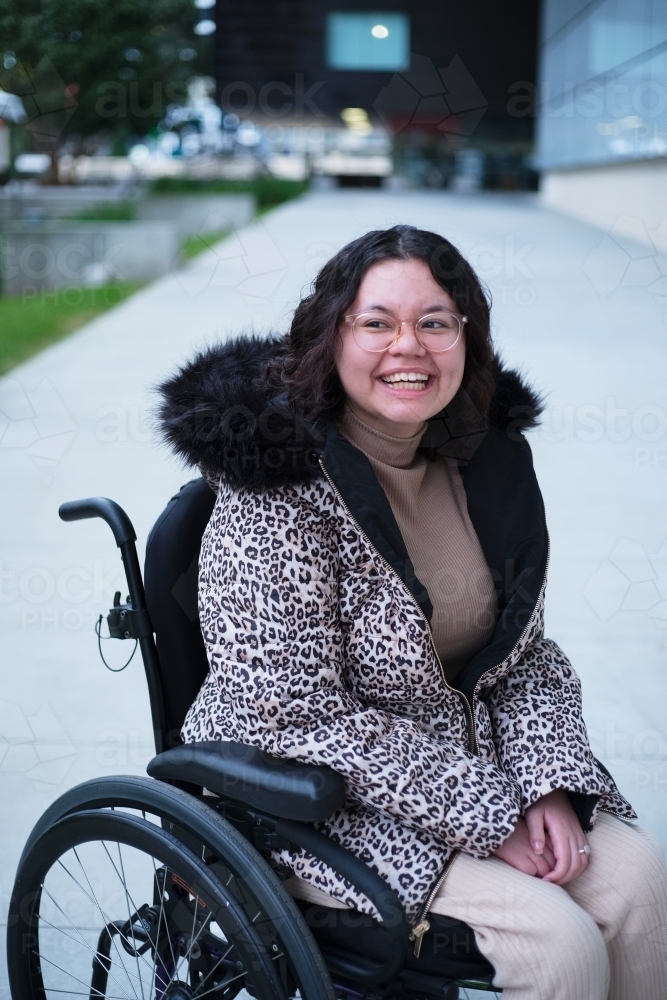 Smiling woman with a disability sitting in a wheelchair outside wearing jacket - Australian Stock Image