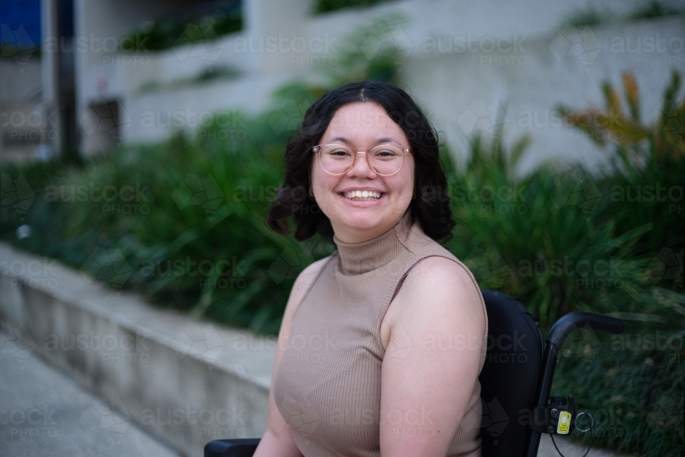 Smiling woman with a disability sitting in a wheelchair outside with grass and plants behind her - Australian Stock Image
