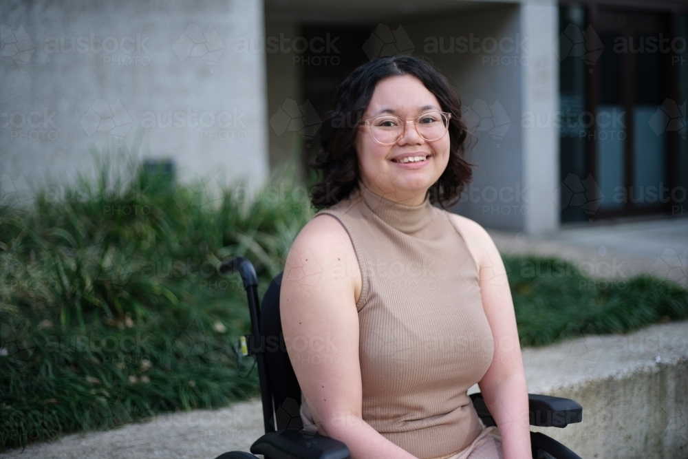 Smiling woman with a disability sitting in a wheelchair outside with grass and plants behind her - Australian Stock Image
