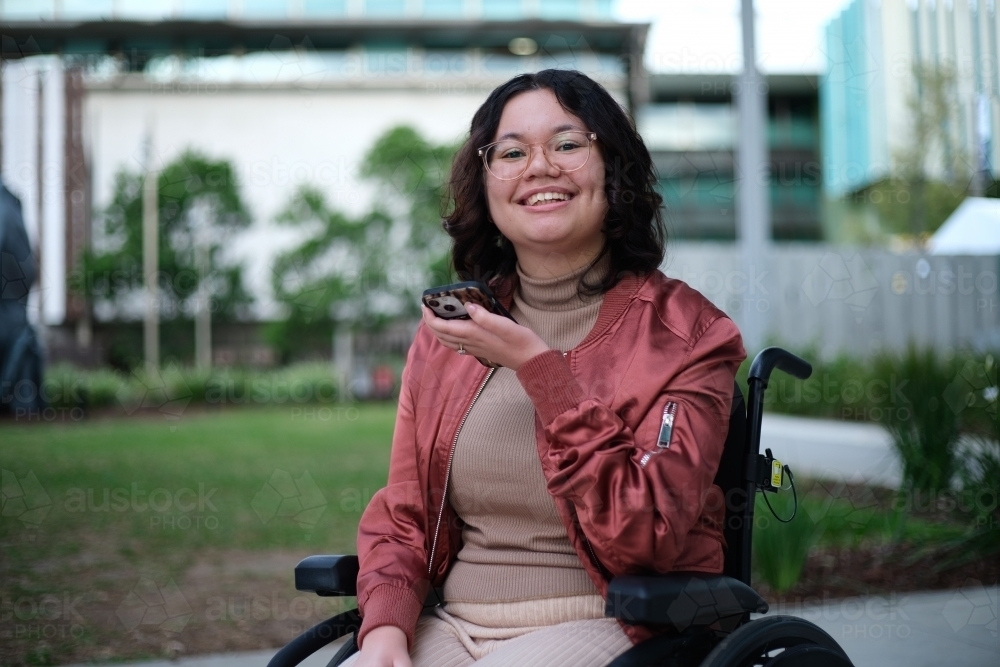 Smiling woman with a disability wearing glasses sitting in a wheel chair holding a mobile phone - Australian Stock Image