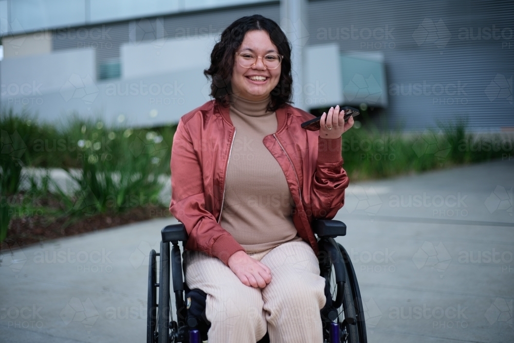 Smiling woman with a disability wearing glasses sitting in a wheel chair holding a mobile phone - Australian Stock Image