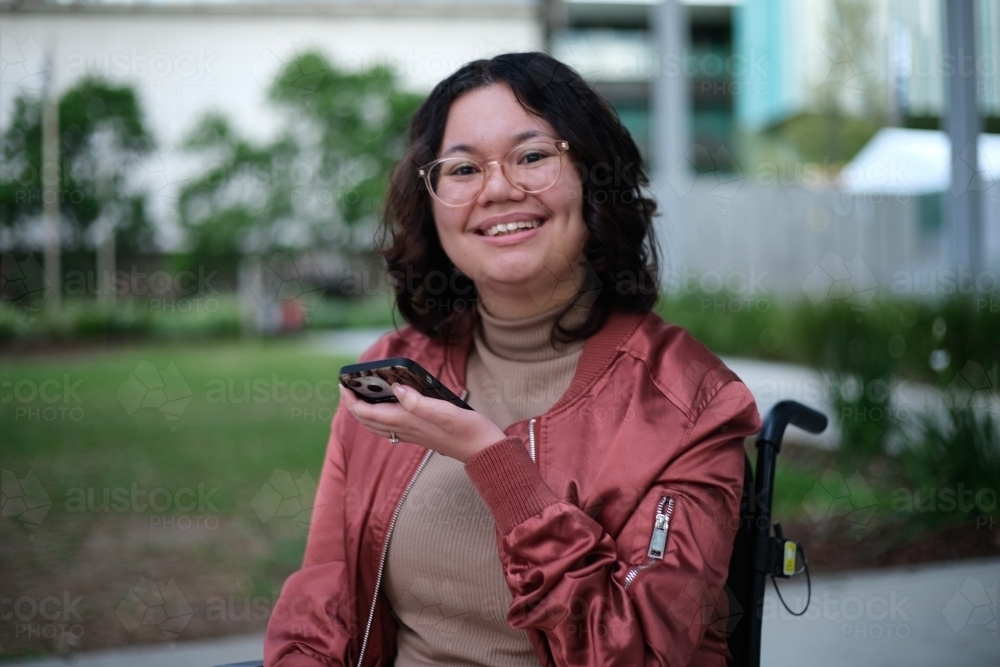 Smiling woman with a disability wearing glasses sitting in a wheel chair holding a mobile phone - Australian Stock Image