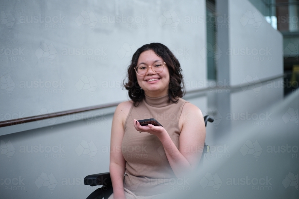 Smiling woman with a disability wearing glasses sitting in a wheel chair holding a mobile phone - Australian Stock Image
