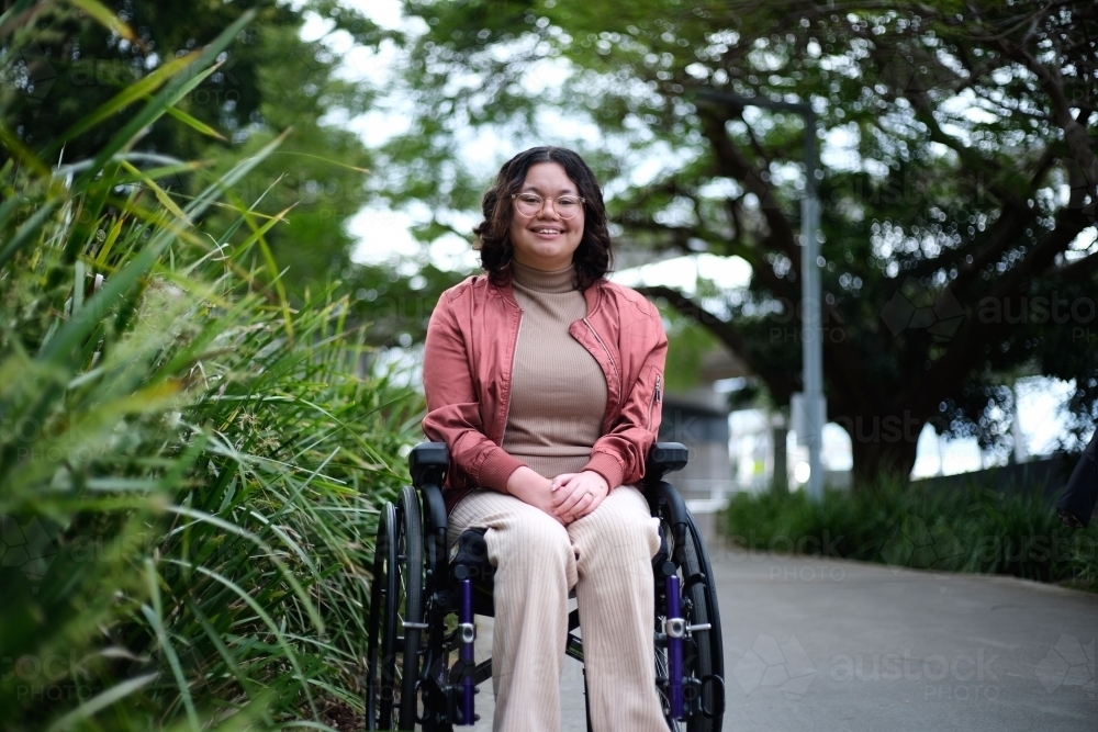 smiling woman with disability sitting in a wheelchair outside next to a tall grass - Australian Stock Image