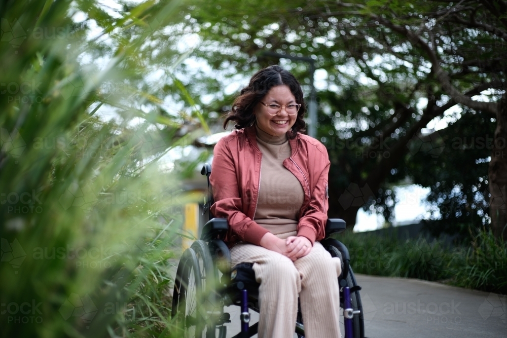 smiling woman with disability sitting in a wheelchair outside next to a tall grass - Australian Stock Image