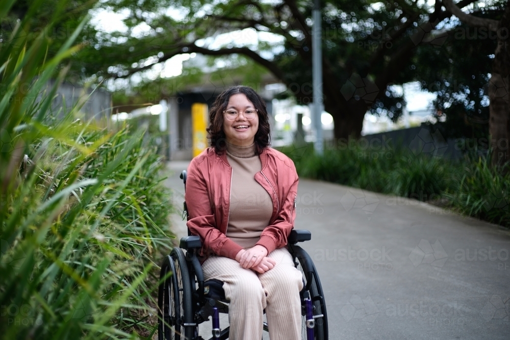 Smiling woman with disability sitting in a wheelchair outside with tall grass and trees around her - Australian Stock Image