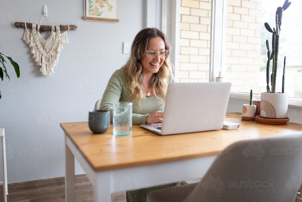 Smiling woman working from home on laptop at kitchen table - Australian Stock Image