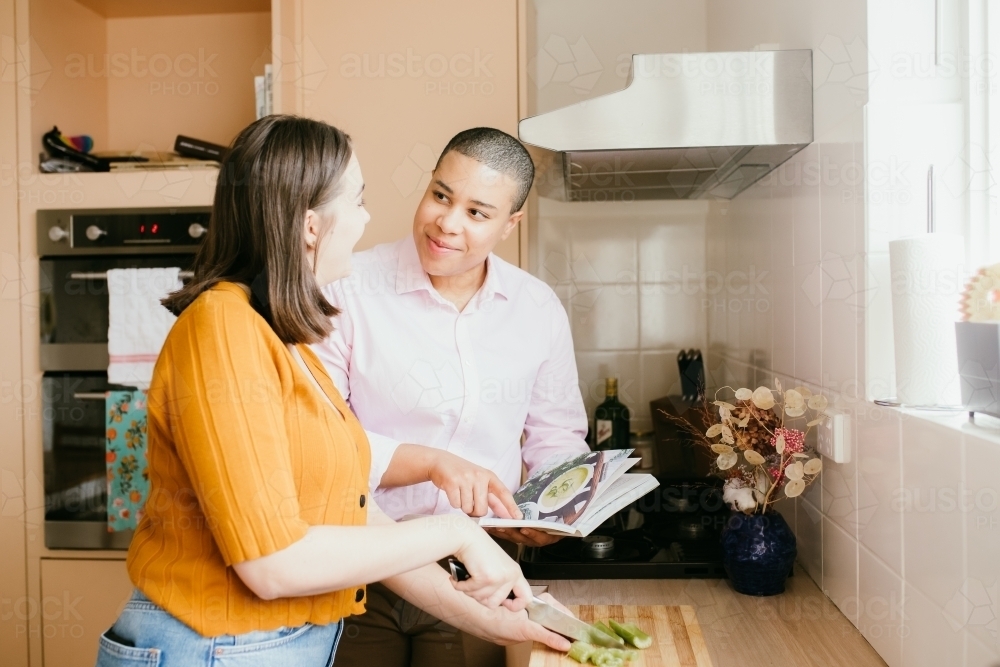Smiling lgbtqi couple with one chopping the leeks and the other one holding a book in the kitchen - Australian Stock Image