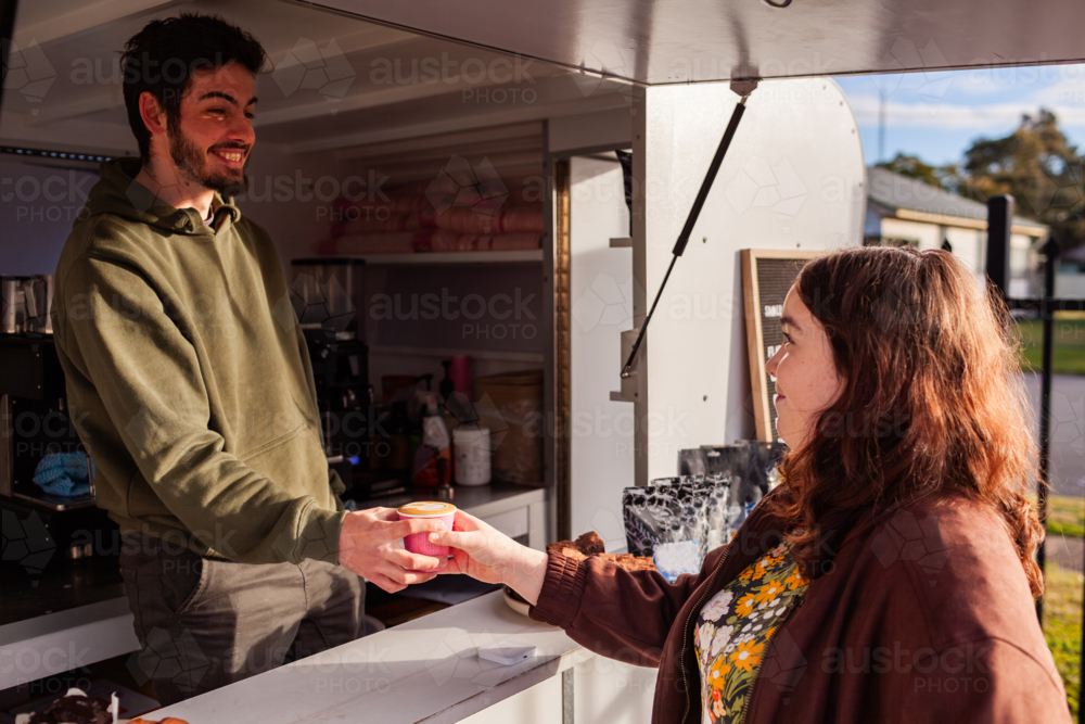 smiling young Aussie barista man handing customer cup from coffee van in morning sunlight - Australian Stock Image
