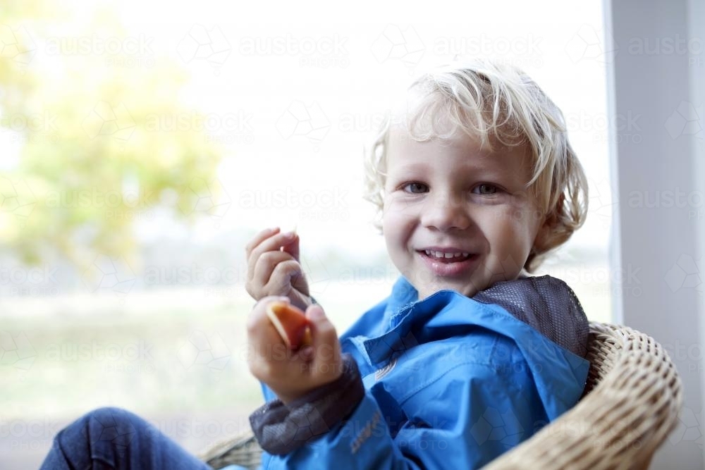 Smiling young boy wearing a blue jacket eating pieces of apple - Australian Stock Image