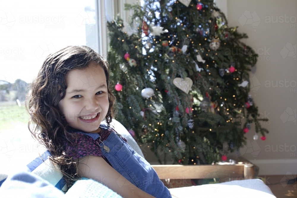 Smiling young girl sitting on chair in front of Christmas tree - Australian Stock Image