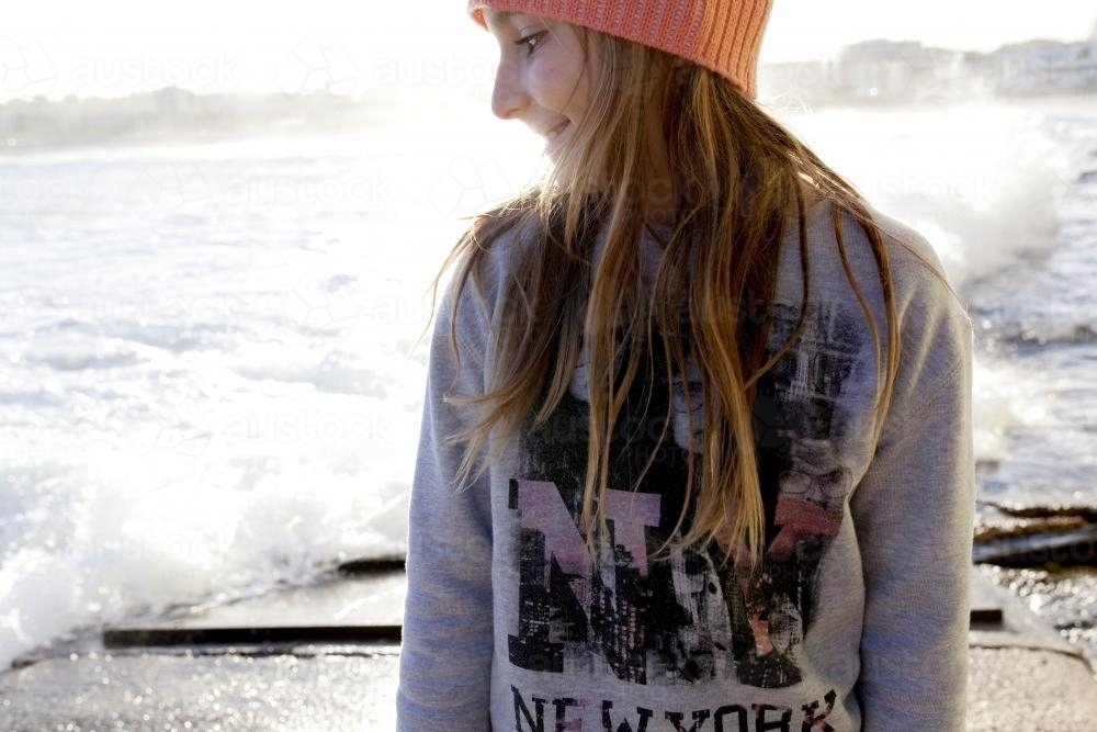 Smiling young girl standing by the ocean looking sideways over her shoulder - Australian Stock Image