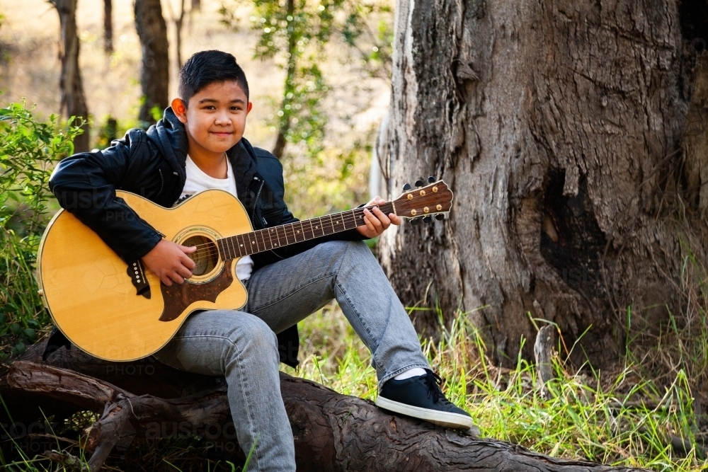 Smiling young guitar player seated on log in bushland - Australian Stock Image