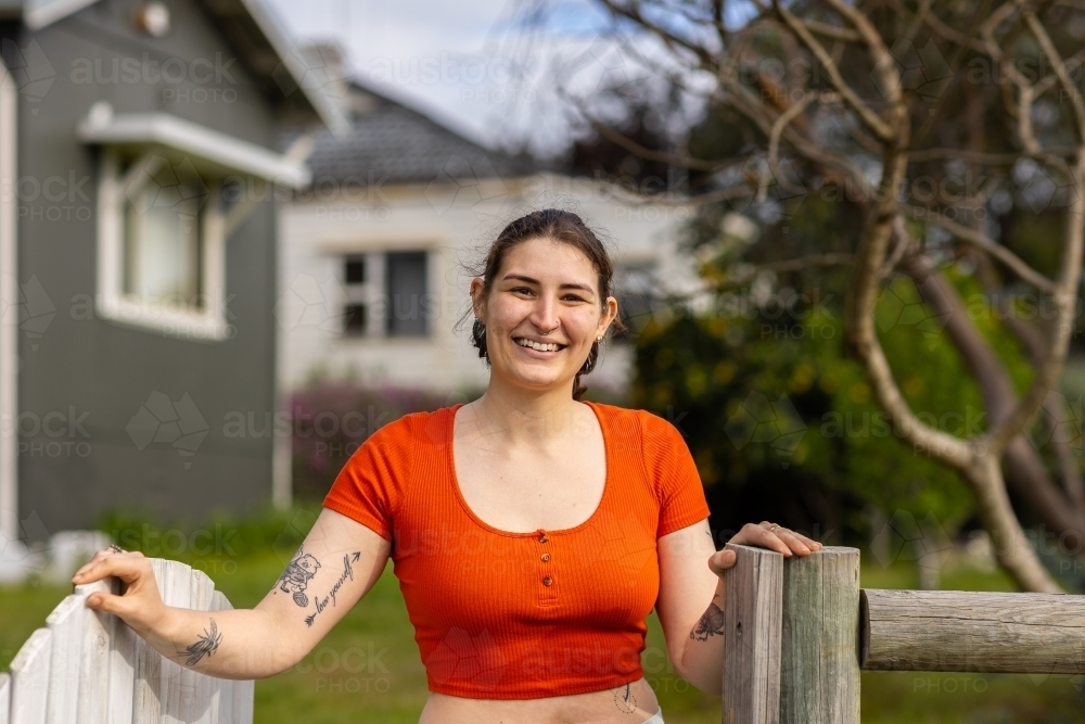 smiling young woman standing outside her home with hand on gatepost - Australian Stock Image