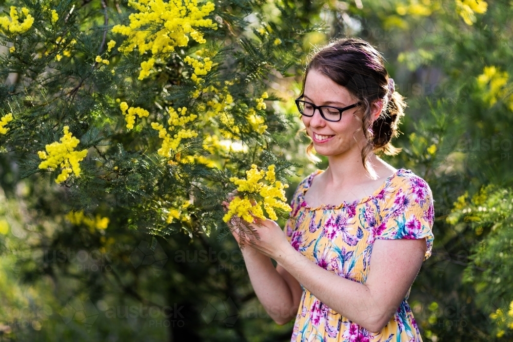 Smiling young woman with glasses standing among leaves and blossoms of wattle bush - Australian Stock Image