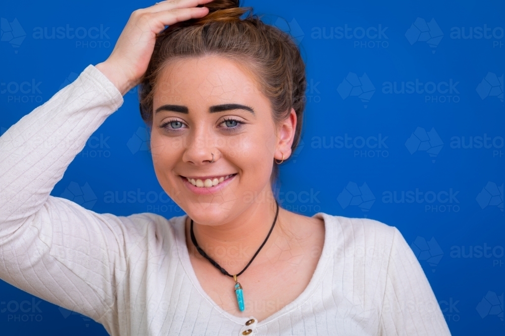 smiling young woman with hair up hand on head against blue background - Australian Stock Image
