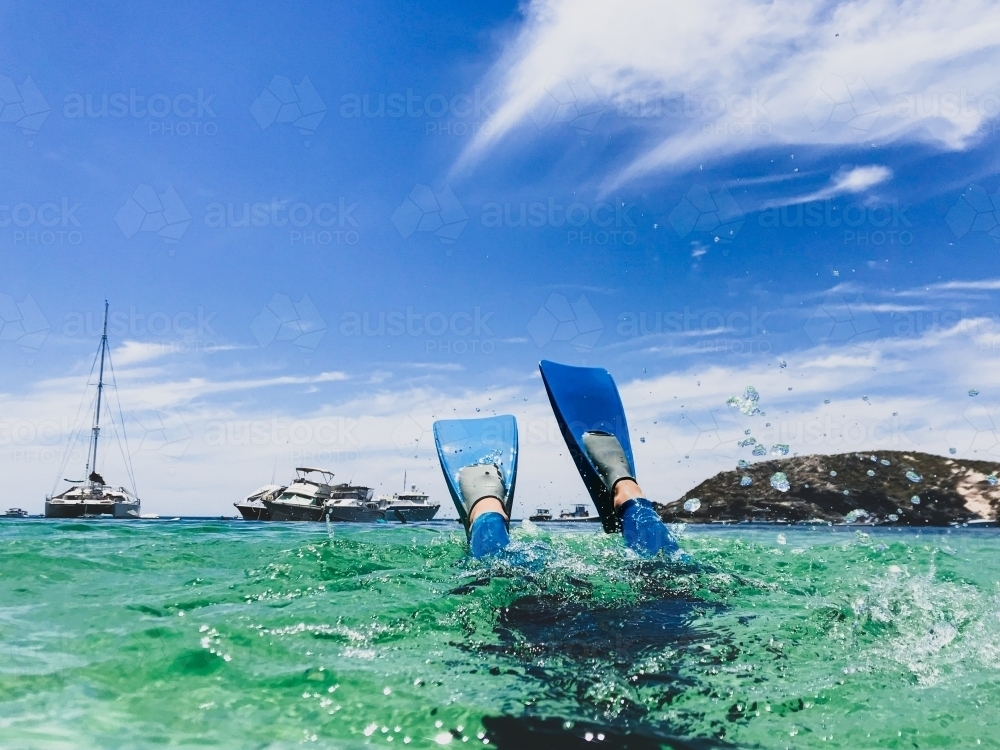 Snorkelers fins splashing above water in clear ocean with boats and land in the background - Australian Stock Image