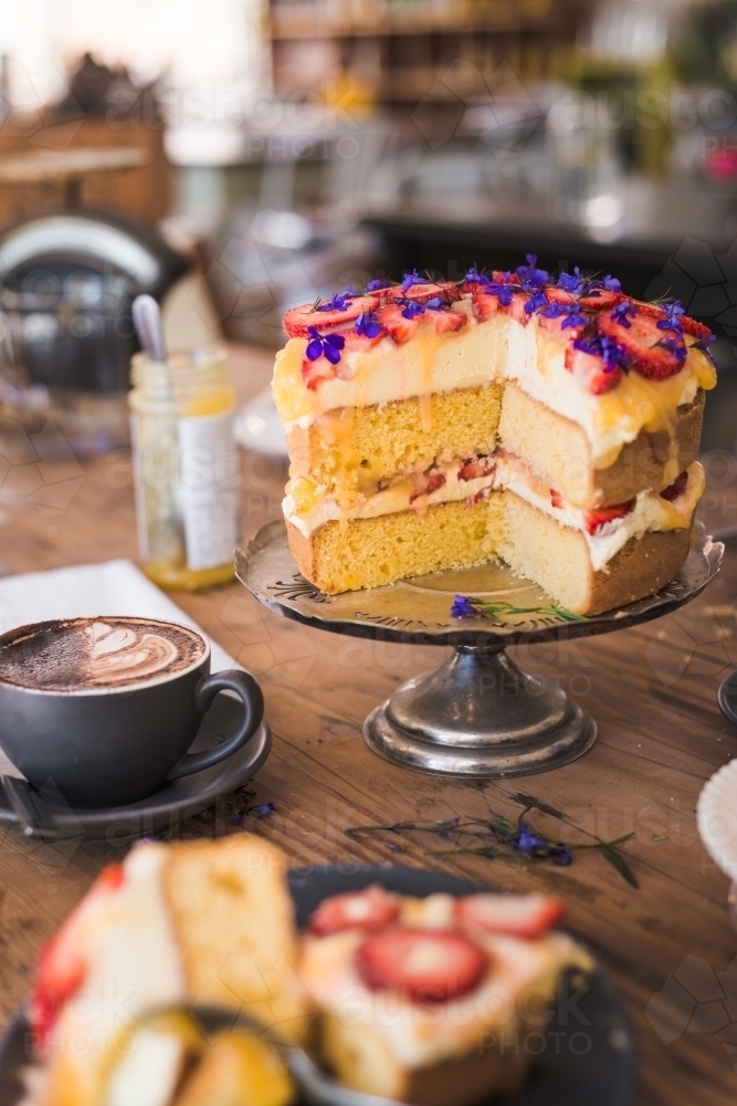 sponge cake with lemon curd, strawberries and edible flowers with pieces cut out, and a coffee - Australian Stock Image