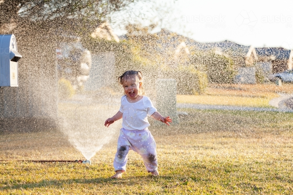 Sprinkler watering grass in front yard of suburban home with little kid running through water spray - Australian Stock Image