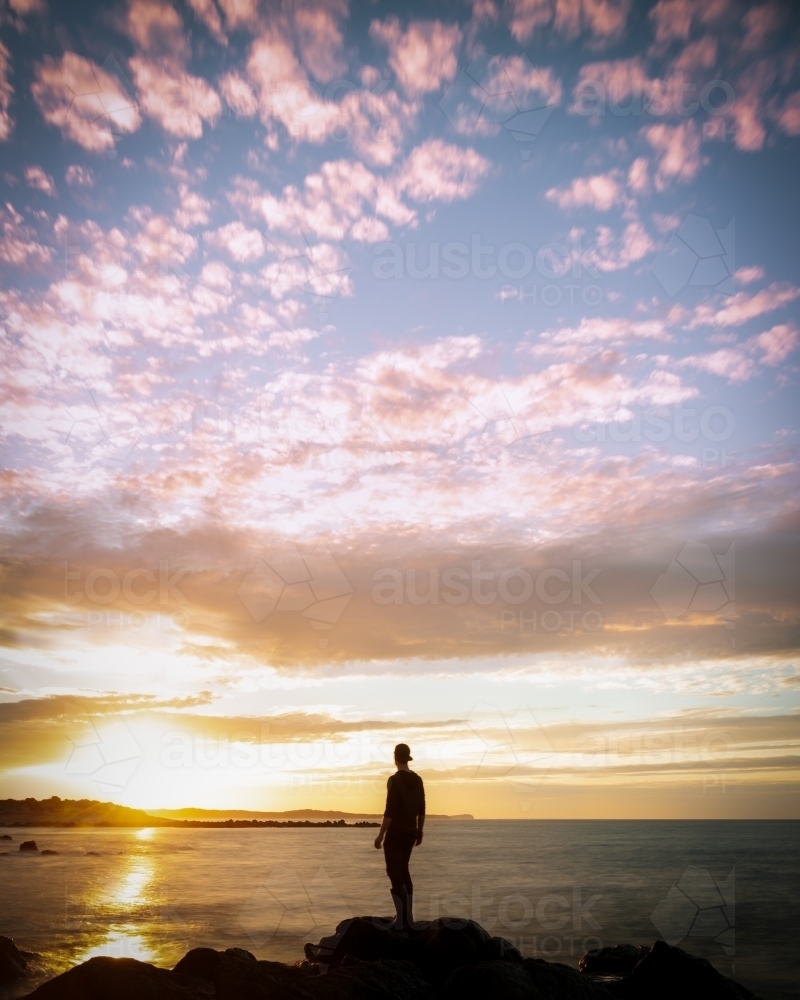Man Standing on the Edge of a Rockpool infront of a Dramatic Sunrise - Australian Stock Image
