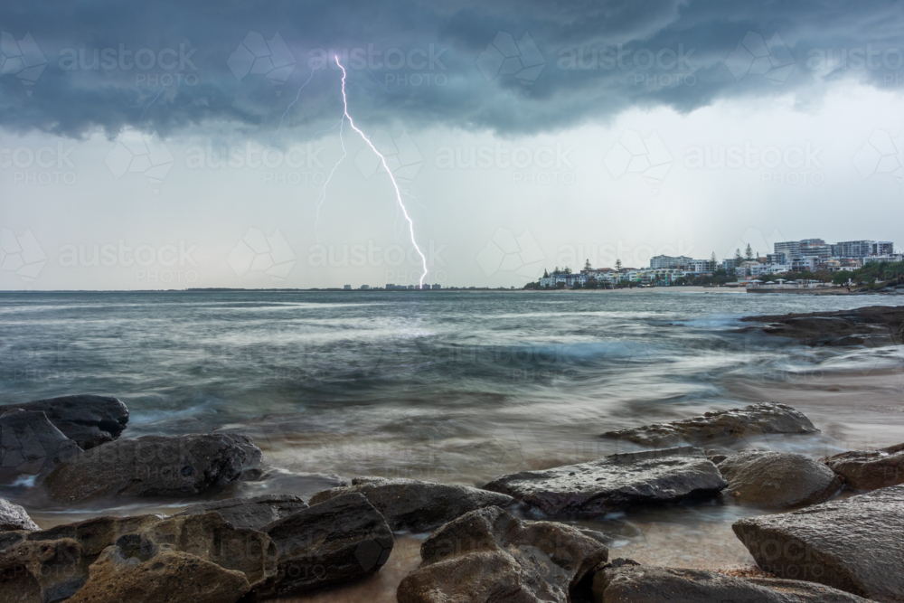 Storm clouds and lightning strike over ocean with rocky shoreline and coastal town in background - Australian Stock Image