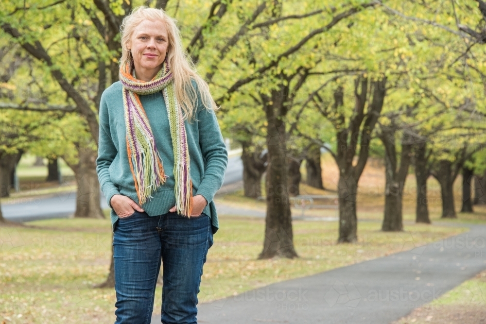 Strawberry blonde woman standing in front of a line of trees in winter wearing a scarf. - Australian Stock Image