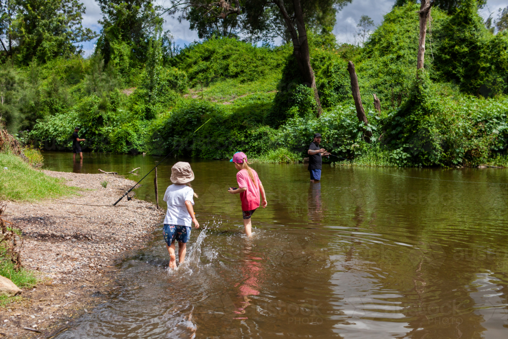 Summertime fun of children playing in river with men fishing in background - Australian Stock Image