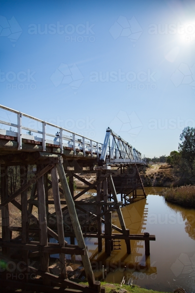 Sun flare over pillars of timber truss road bridge above creek - Australian Stock Image