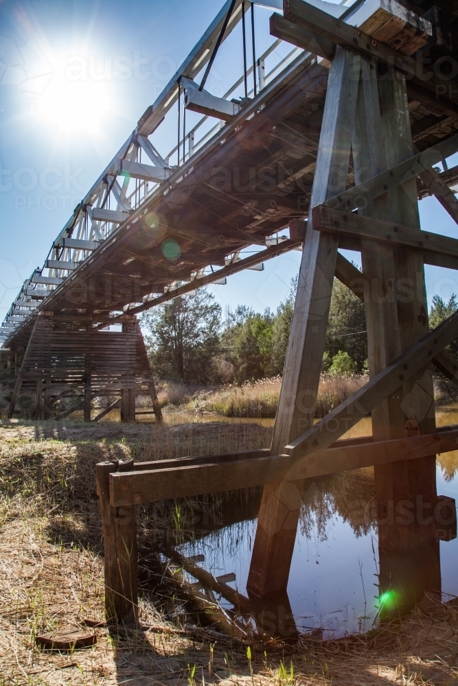 Sun flare over pillars of timber truss road bridge above creek - Australian Stock Image