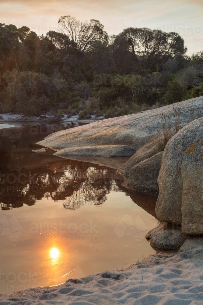 Sun reflection in still water surrounded by boulders on beach - Australian Stock Image