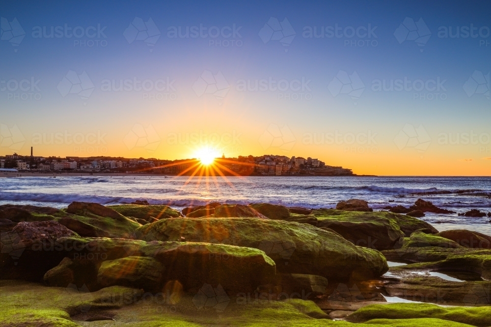 Sun rising over moss covered rocks along coastline with blue sky - Australian Stock Image