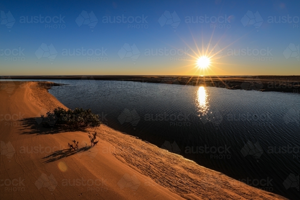 Sun rising through blue sky over ocean channel and sandy coastline - Australian Stock Image