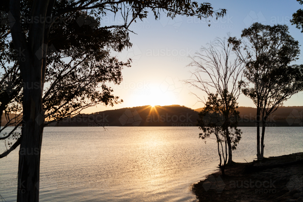 Sun setting across lake waters with silhouetted gum trees in foreground - Australian Stock Image
