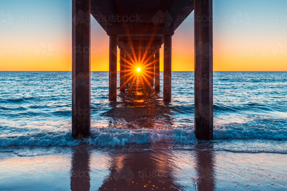 Sun shining through Brighton Jetty pylons while viewed from the beach at sunset time - Australian Stock Image