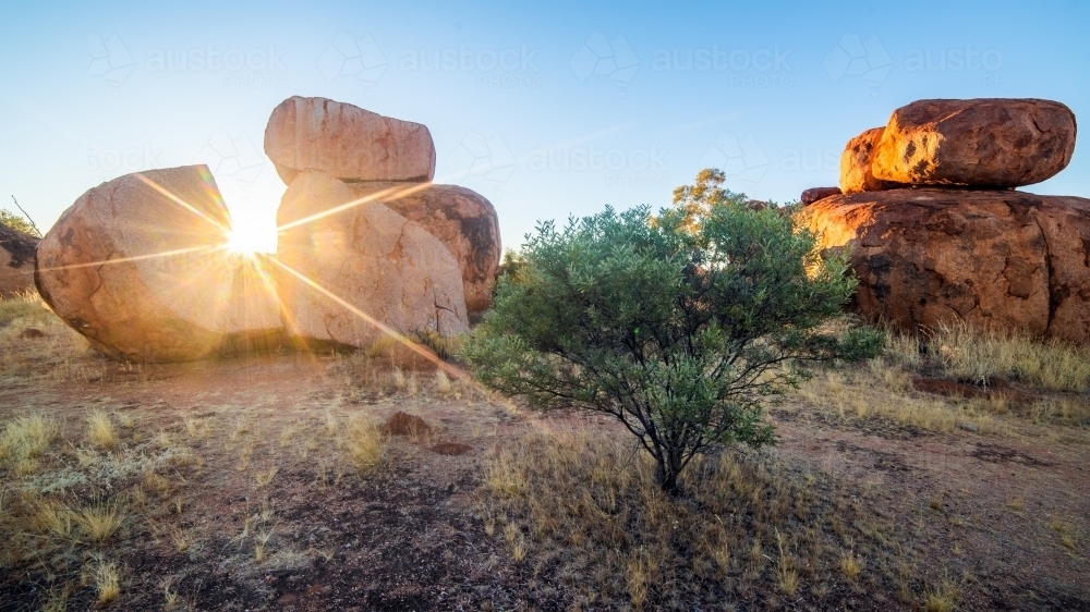 Sun shining through large boulder at sunrise - Australian Stock Image