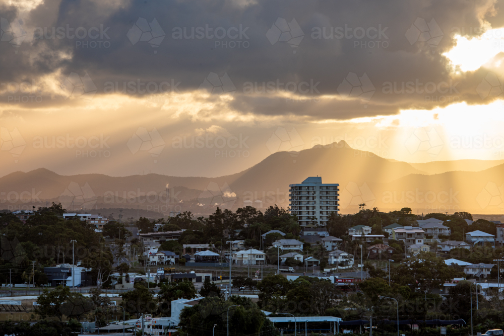 Sunbeams breaking through clouds over mountain silhouette and coastal town buildings at sunset - Australian Stock Image