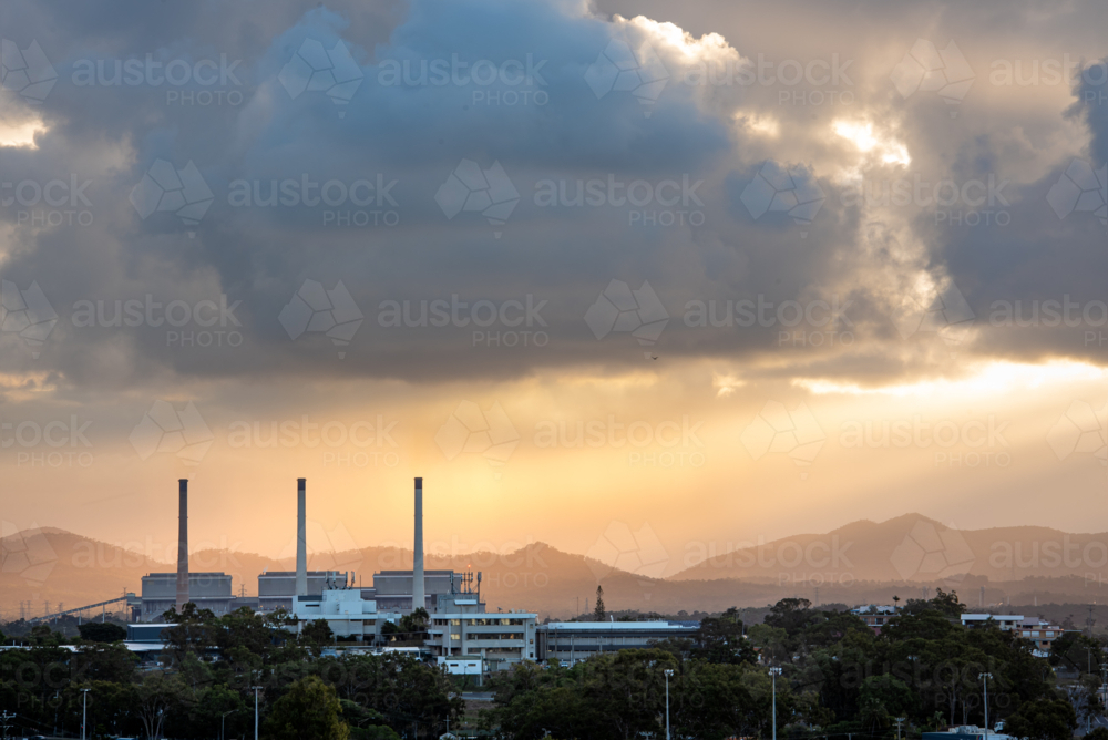 Sunlight breaking through storm clouds over city landscape and industrial smokestacks at sunset - Australian Stock Image