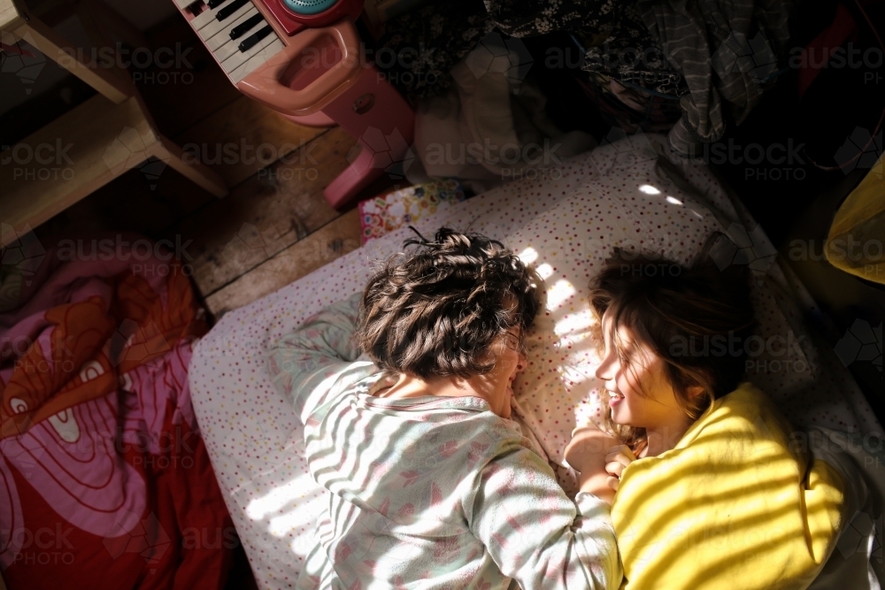 Sunlight falling on two girls smiling together on bed on the floor - Australian Stock Image