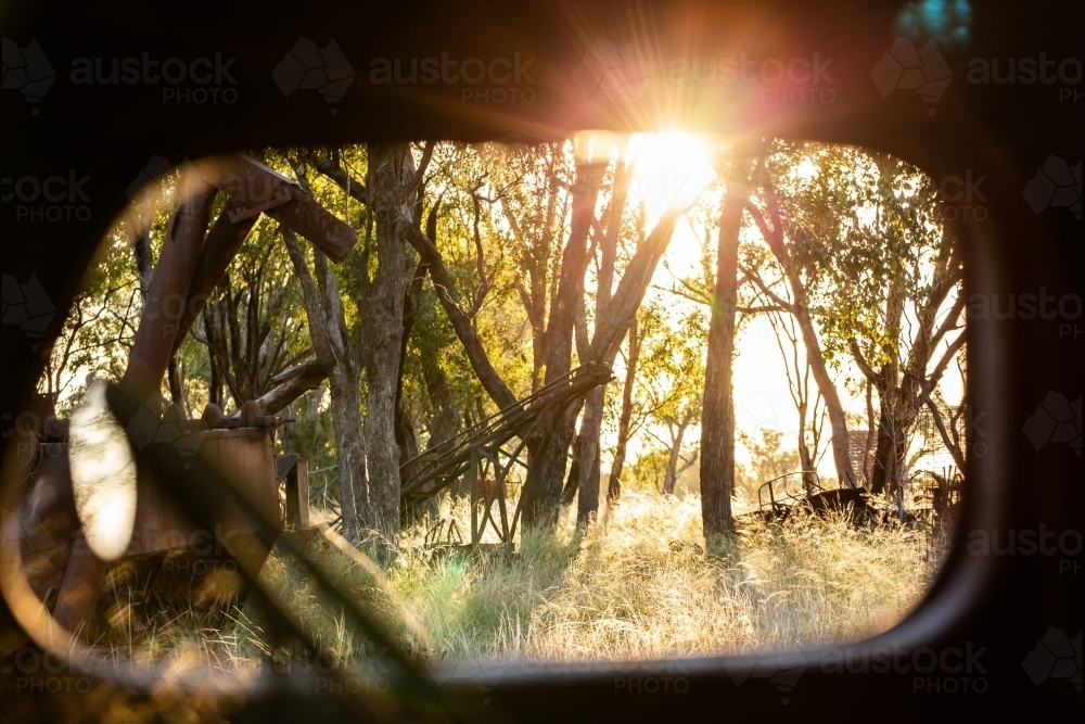 Sunlight flare through trees in farm machinery junkyard seen through rusted out window of vehicle - Australian Stock Image