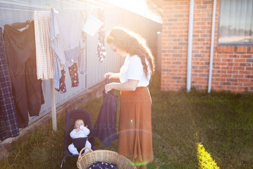 Sunlight over young mother getting washing in with baby in bouncer - Australian Stock Image